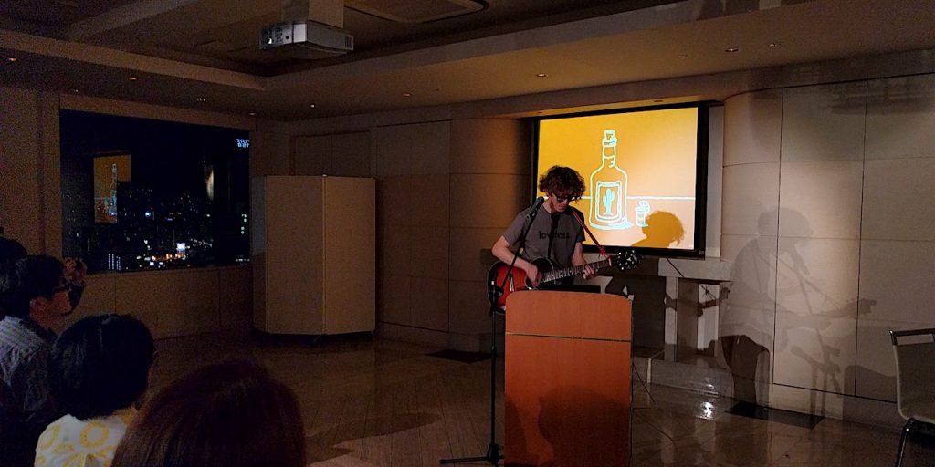自作のスライドを前にギターを引きながら歌う澤田さんの写真