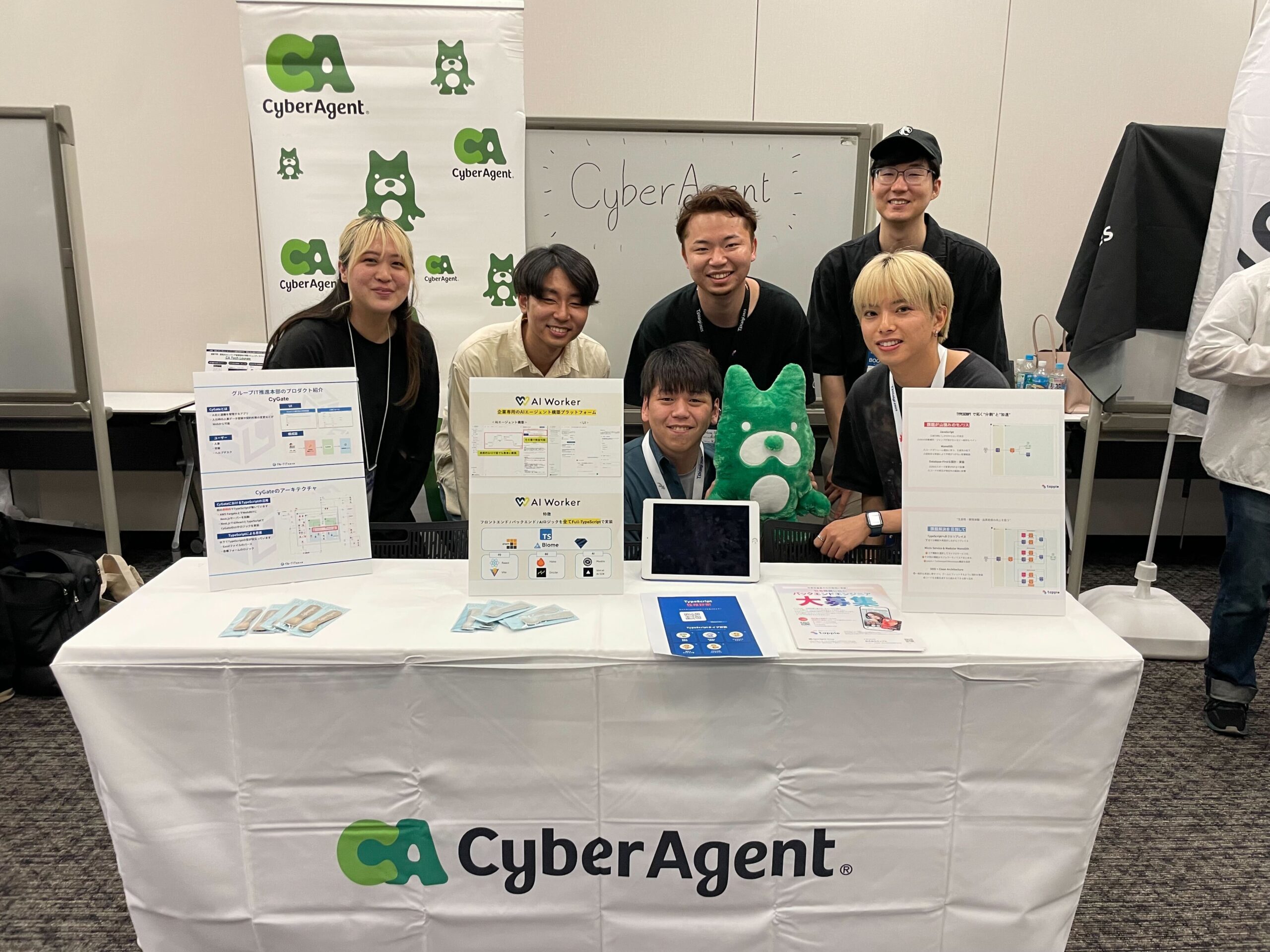 CyberAgentのロゴが掲げられたブースで、社員6名が笑顔で並んでポーズを取っている。テーブルにはプロダクトの紹介資料やタブレット、チラシが展示されており、中央には緑色のアベマくんのぬいぐるみも置かれている。背景にはCyberAgentのロゴバナーとホワイトボードがある。