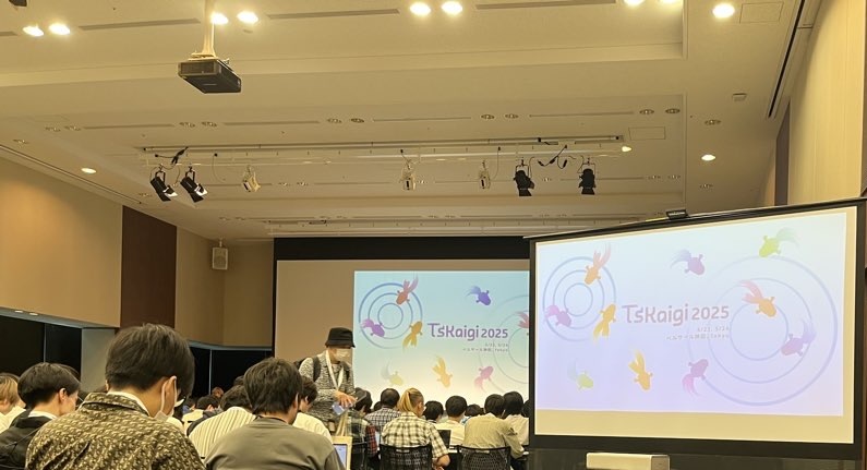 カンファレンス会場の室内で、多数の参加者が着席している。正面のスクリーンとサブスクリーンには「TSKaigi 2025」のロゴとカラフルな金魚のイラストが表示されている。